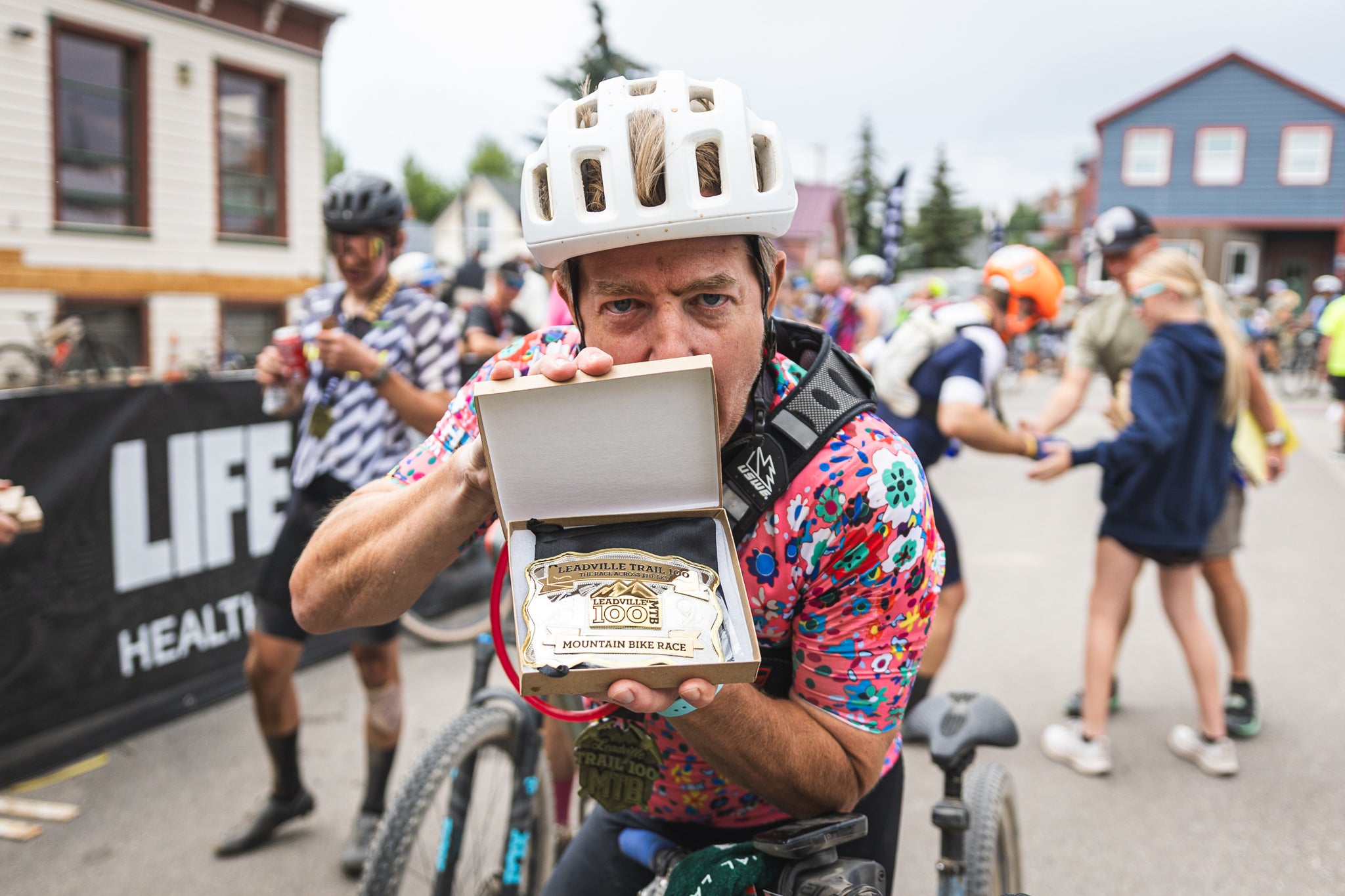 Rack Rabbit founder Edward Wimmer, wearing a white helmet and colorful floral jersey, proudly displays his Leadville Trail 100 MTB finisher belt buckle in its presentation box at the race finish line, surrounded by fellow cyclists.
